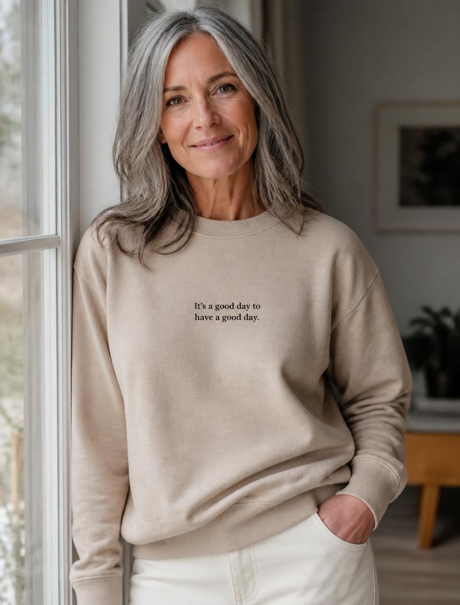 Person in a sand crewneck with a small printed line on the chest, standing by a window